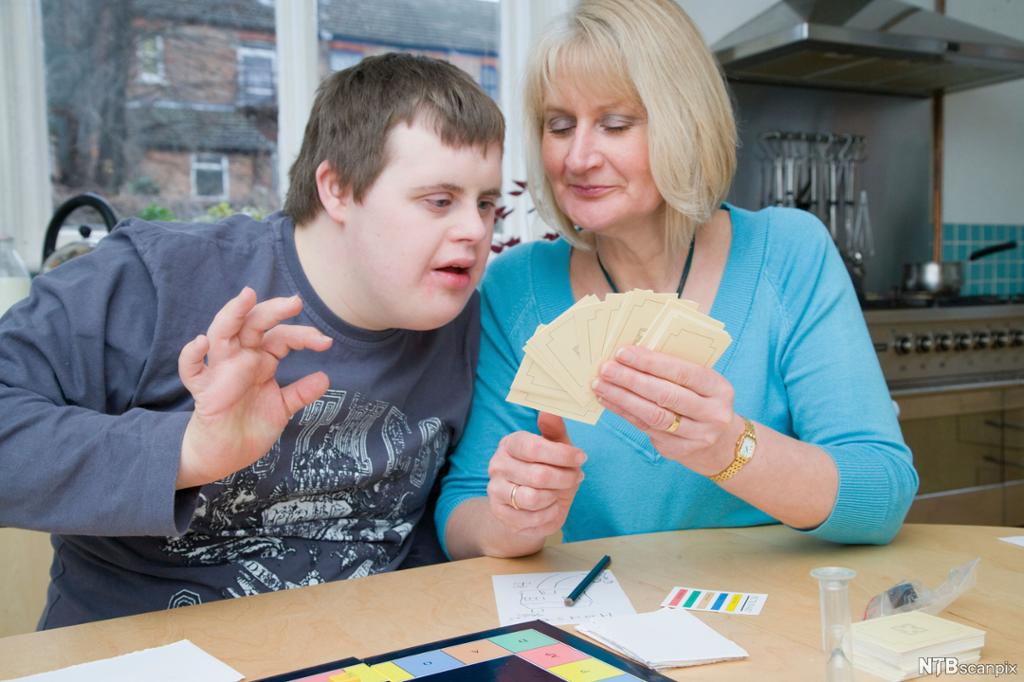 Ung mann med Downs syndrom spiller spill. En kvinne sitter ved siden av ham og holder en bunke kort som han ser engasjert på. Det er spillebrett, timeglass, blyant og papir på bordet foran dem. Foto. 
