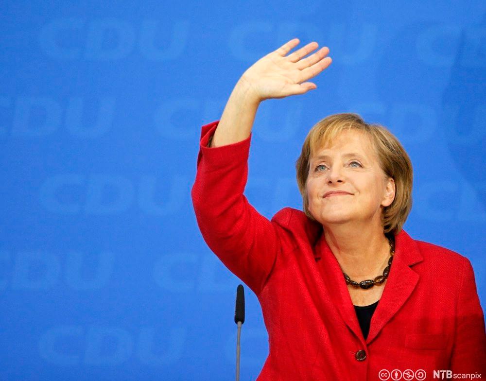 Den tyske politikeren Angela Merkel på et partimøte. Hun har på seg en rød blazer over en sort genser, og et halskjede satt sammen av store, mørke, avlange steiner. Hun ser opp, vinker og smiler. Foto.