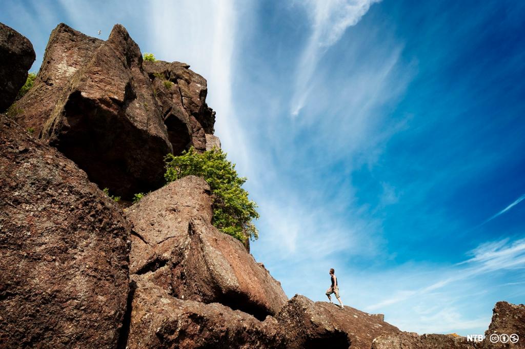 Photo: Steep mountain side, man standing at the bottom, blue sky above. 
