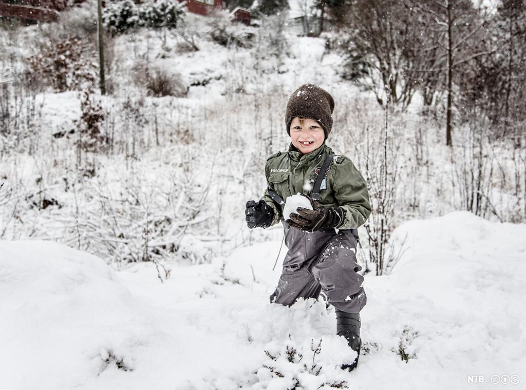 En smilende gutt ute i snøen med en snøball i hånda. Foto.