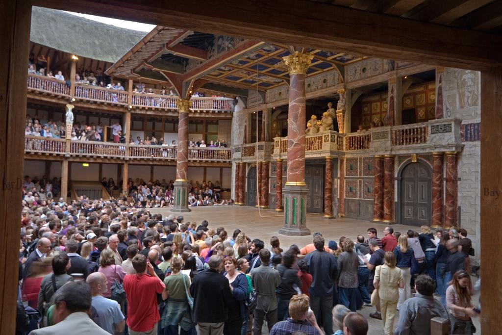 A photo taken inside The Globe Theatre in London. People are waiting for the play to start. Most people are standing in front of the stage. 