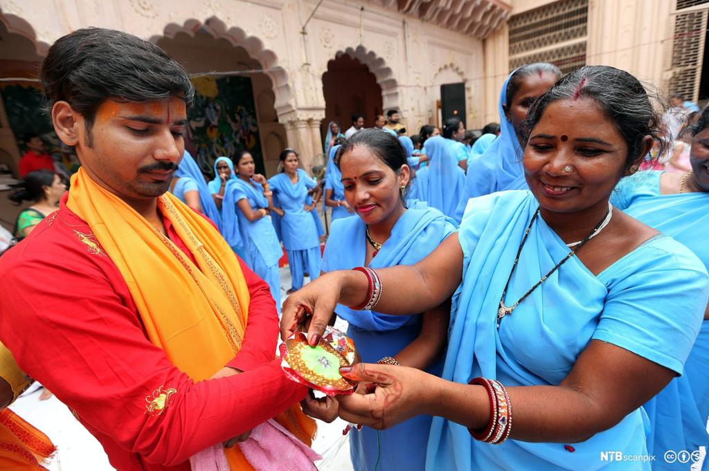 En hindukvinne i blå drakt knyter et hellig bånd på en mann fra en høyere kaste kledd i rødt og orange. Det er mange kvinner i bakgrunnen. Foto. 