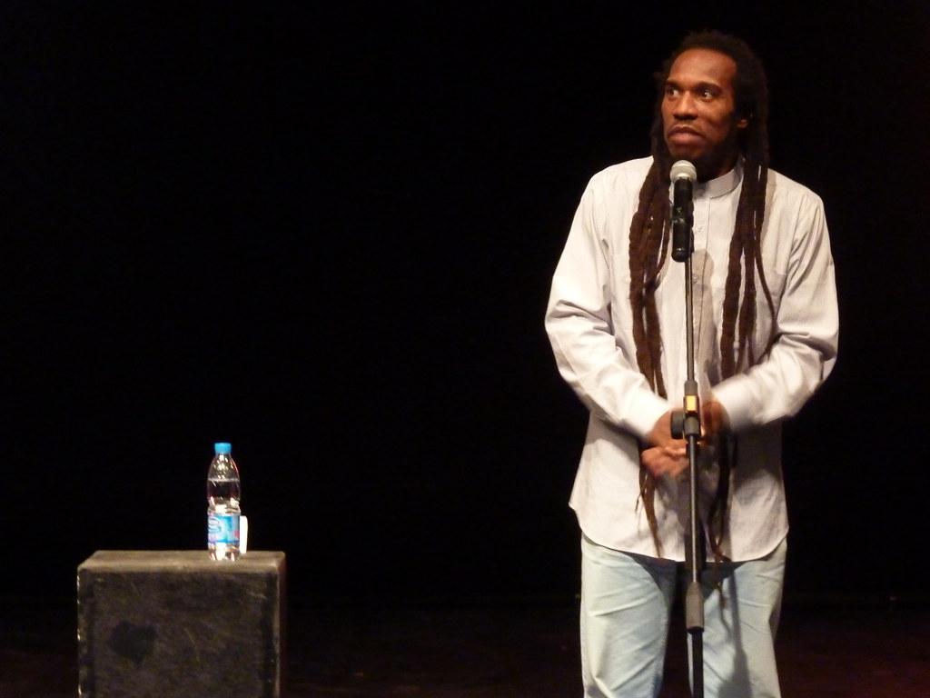 A photo of British dub poet Benjamin Zephaniah on stage during one of his performances. He's a black man with long dreadlocks, dressed in a white shirt and jeans. 