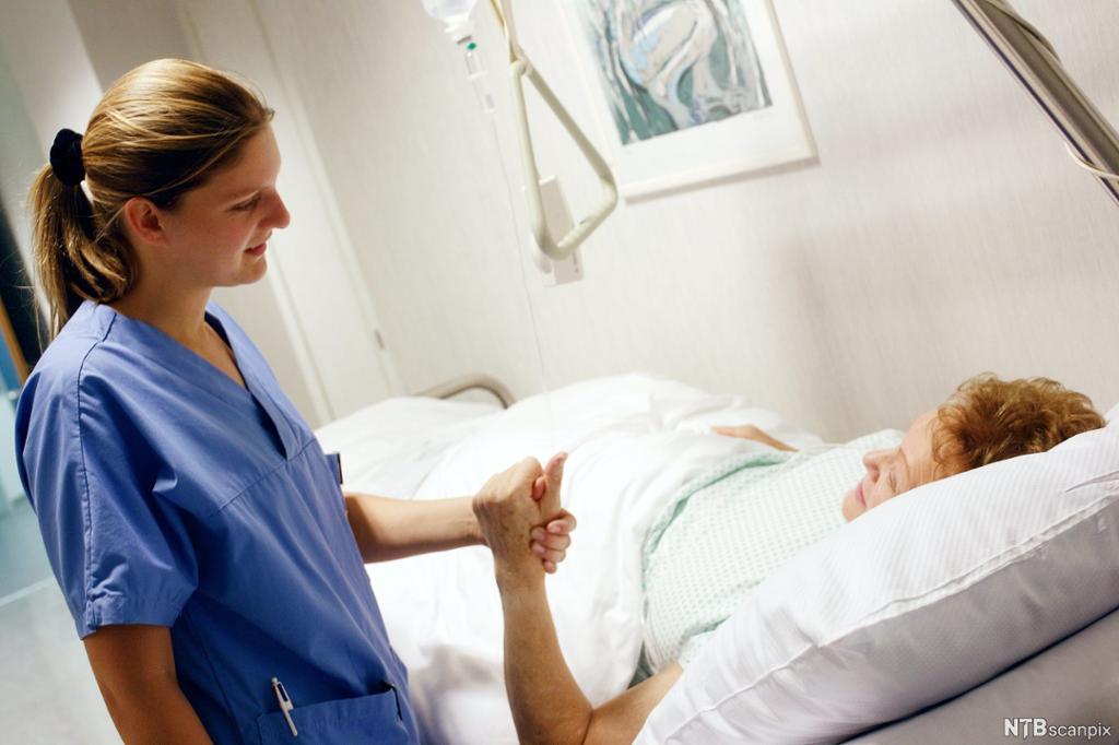 En helsefagarbeider holder hånden til en pasient som ligger i sykehusseng. Foto.