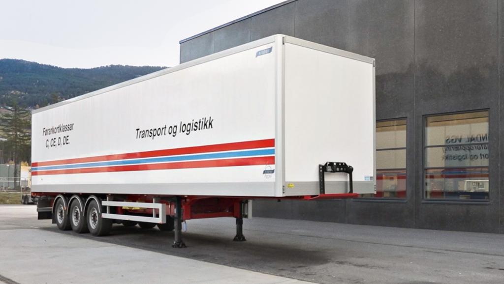 En semitrailer uten trekkvogn står parkert ved et grått industribygg. På traileren står det «Førerkortklasser C, CE, D, DE» og «Transport og logistikk». Nederst er den dekorert med en påmalt stripe i fargene til det norske flagget. Foto.