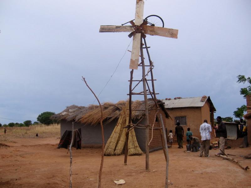 Simple windmill primarily built in wood. Huts and people in the background. Photo.