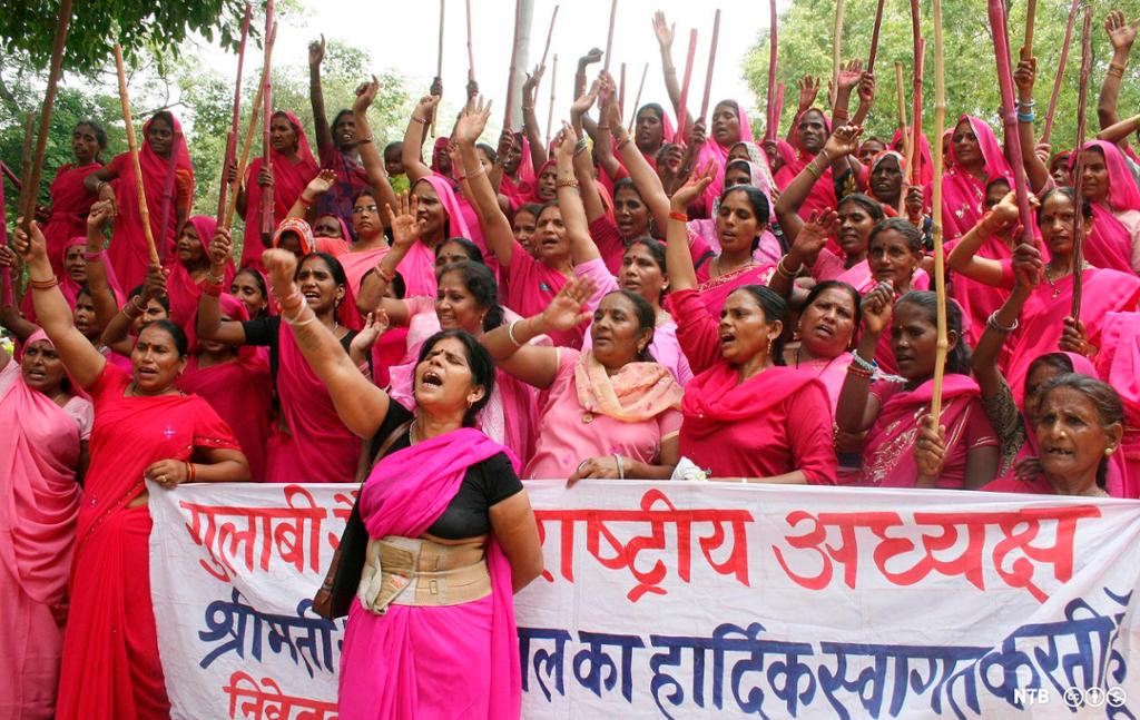 Mange indiske kvinner iført rosa sari holder en hånd i været og roper slagord. De holder opp et stort banner med indisk skrift. Foto.