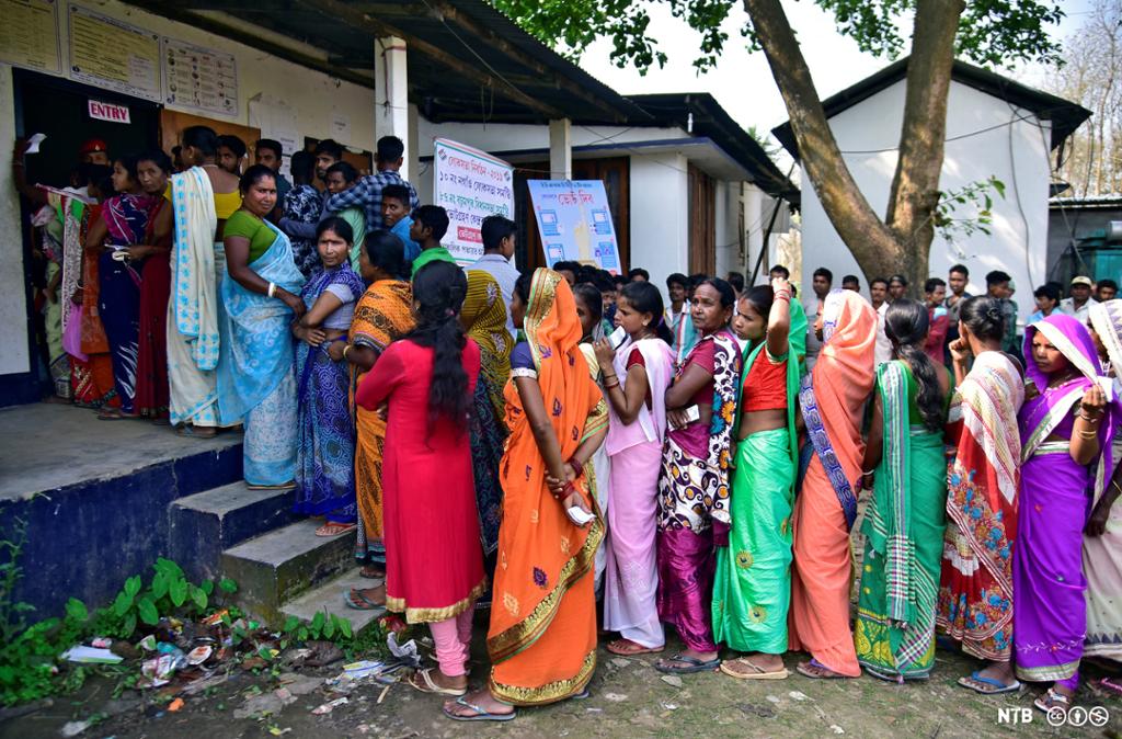 A number of women standing in line outside a polling station, waiting to cast their vote during an Indian election. They are all dressed in colourful saris. The men have lined up in the background in a different queue. Photo.