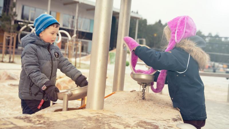 Gutt i blå lue leker med jente i rosa lue. De er utendørs i med lekeapparater og leker med sand. Foto.