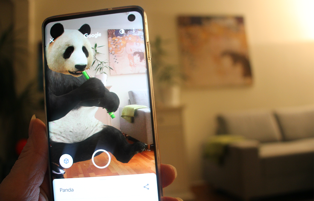 Ei hånd holder en mobil foran en sofa på ei stue. På mobilen er det en panda som sitter på gulvet, ved den samme sofaen. Foto.
