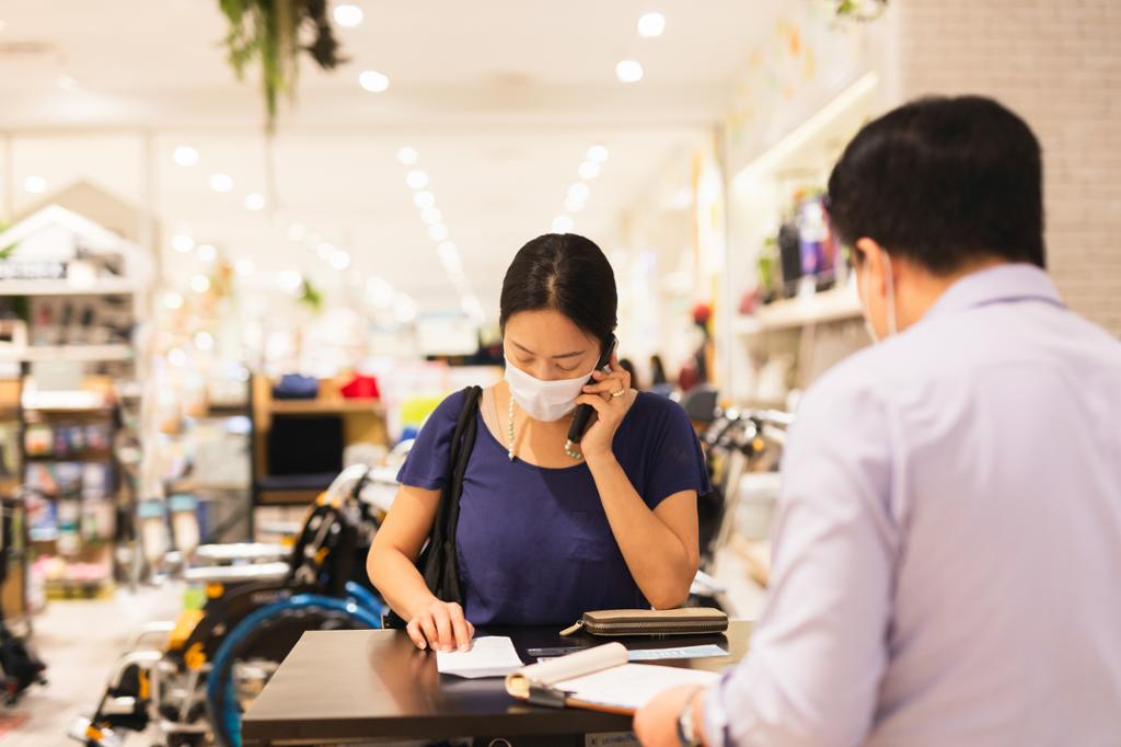 Kvinne med munnbind snakker i telefonen mens hun står ovenfor en mann i kassa i en butikk. Foto. 