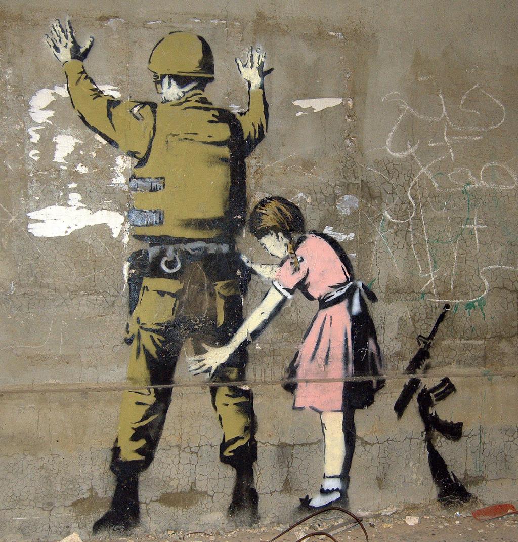 A soldier stands with his hands in the air and his back to the side. A young girl in a pink dress conducts a body search. Photo.