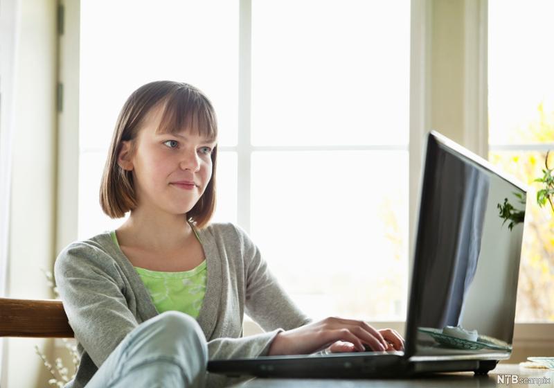 Jente jobber på datamaskin. Hun skriver på tastaturet til laptop-en og ser fornøyd inn i skjermen. Foto.