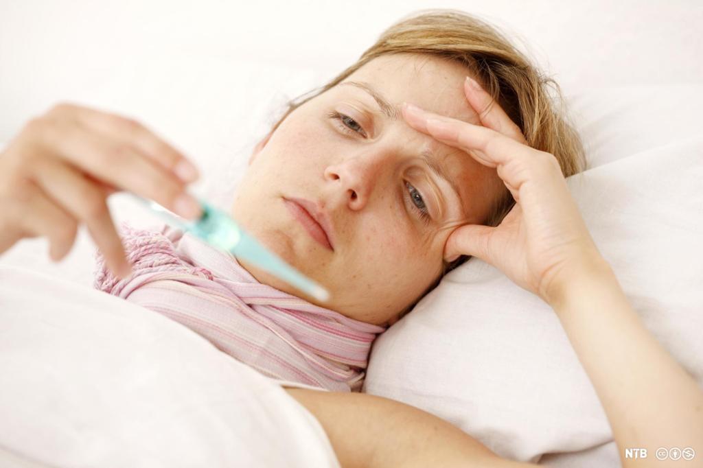 Ung kvinne ligg i senga med eit digitalt termometer i handa. Foto.