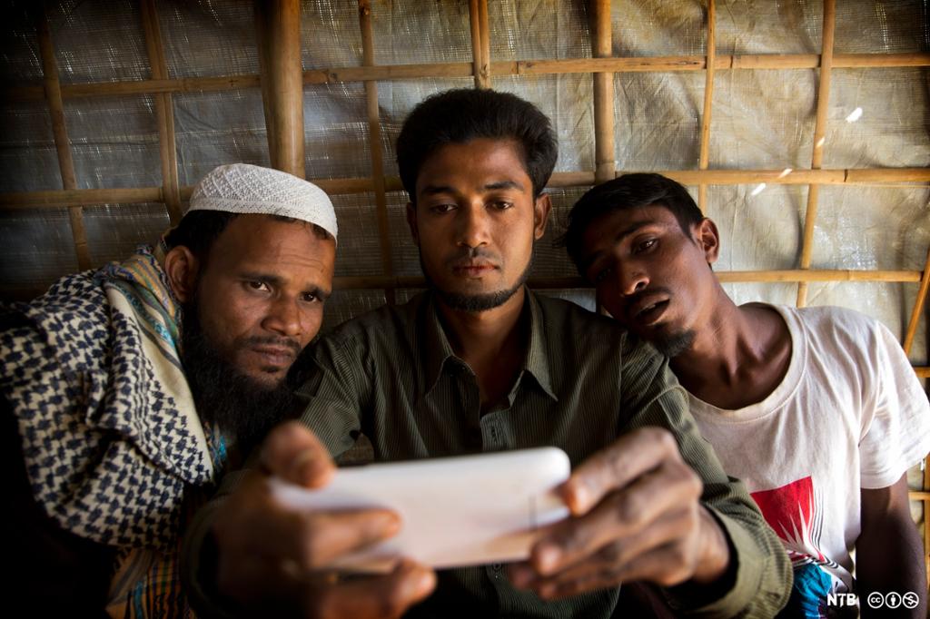 Tre rohingyaer, ein av dei med kufi på hovudet, studerer ein mobilskjerm. Foto.