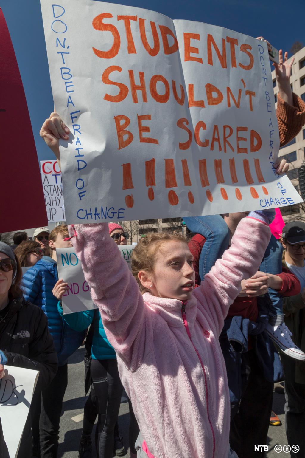 A young girl is participating in the March for Our Lives demonstration in Washington DC in 2018. She's holding a poster over her head saying "Students shouldn't be scared". 