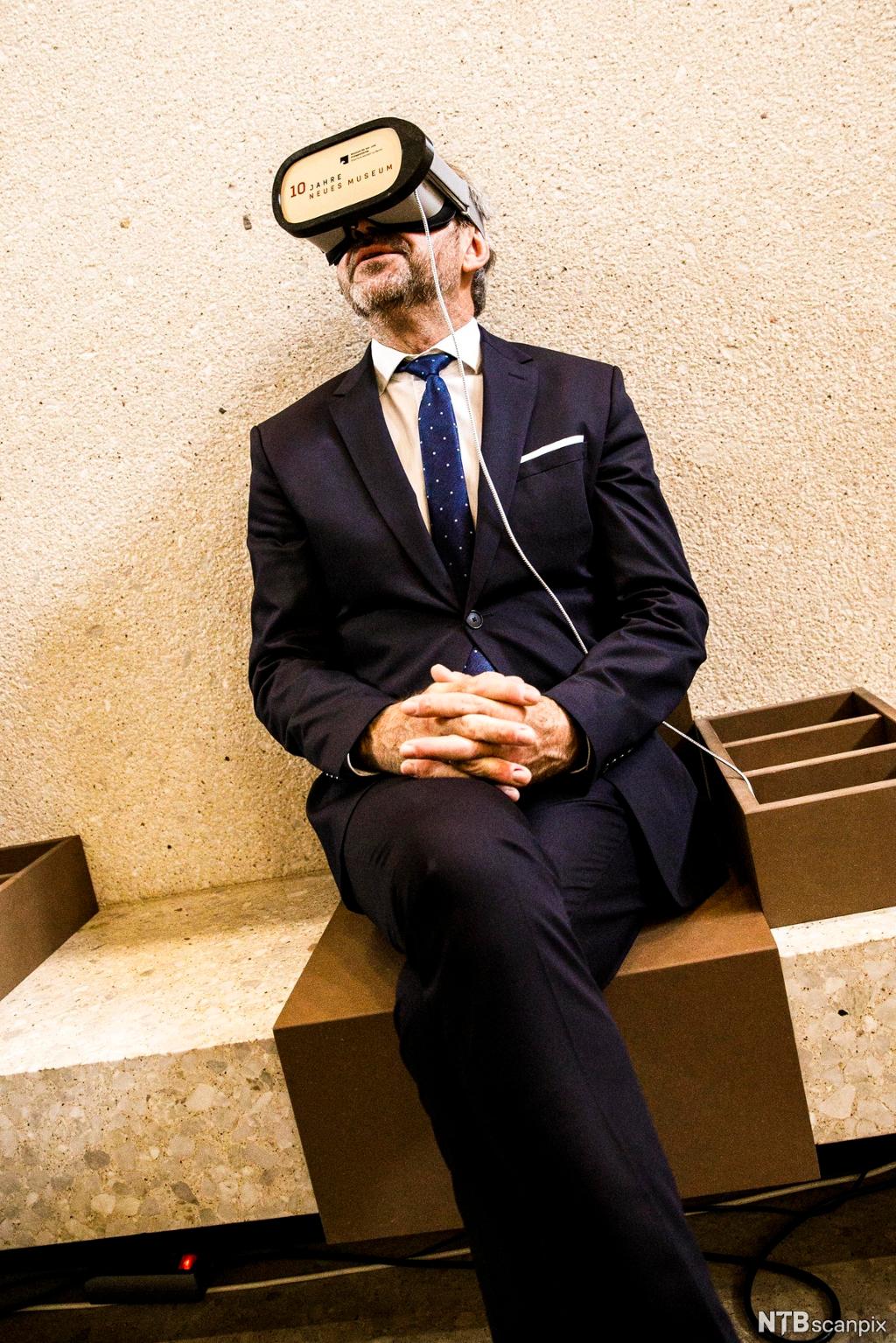 Mann med VR-briller sitter på en benk. Foto.