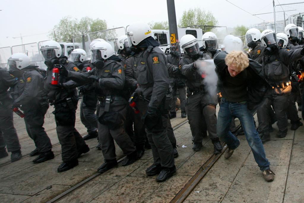 Ei gruppe politifolk under ein demonstrasjon. Dei er utstyrte med tåregass og har på seg hjelm og gassmaske. Ein av dei sprøytar tåregass mot ein demonstrant. Foto.