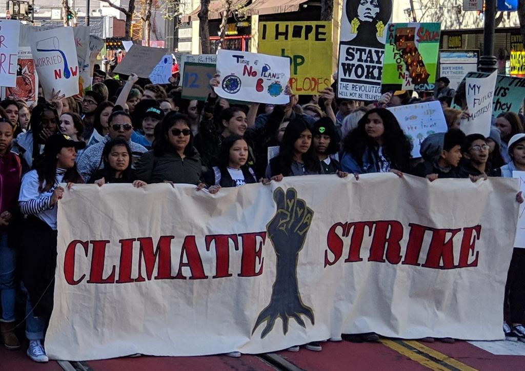 Large banner with a clenched fist with roots and the words Climate Strike, many young people walking in protest behind the banner. 