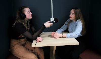 To jenter sitter ved et bord. Jenta til venstre har øreklokker og lydopptaker og lytter til jente til høyre som snakker. Foto.
