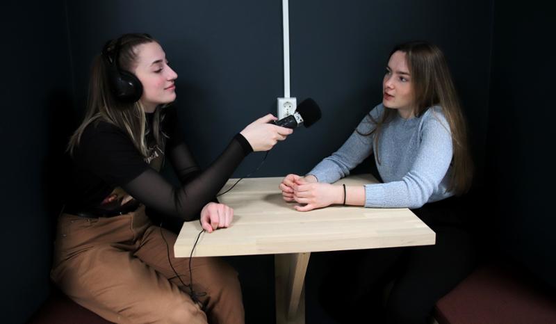 To jenter sitter ved et bord. Jenta til venstre har øreklokker og lydopptaker og lytter til jenta til høyre, som snakker. Foto.