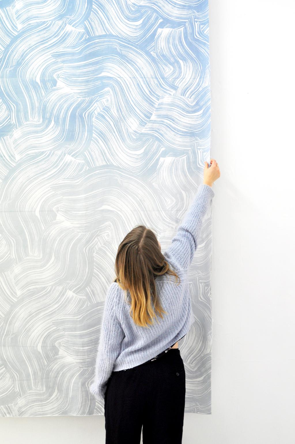 Kvinne strekker seg og kjenner på et tekstiltrykk med bølgede linjer som henger på en vegg. Foto.