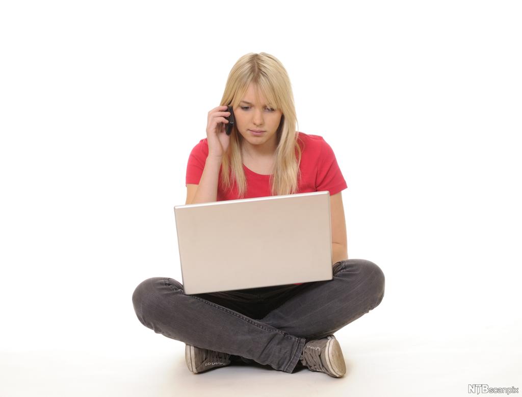 Foto av kvinne som sit med beina i kors med PC mens ho snakkar i ein mobiltelefon. 