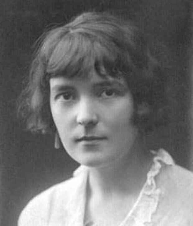 Photo: Black and white portrait of Katherine Mansfield. We see a woman with dark hair, and dark eyes. She is wearing a white blouse with a frilly collar. She looks serious. 