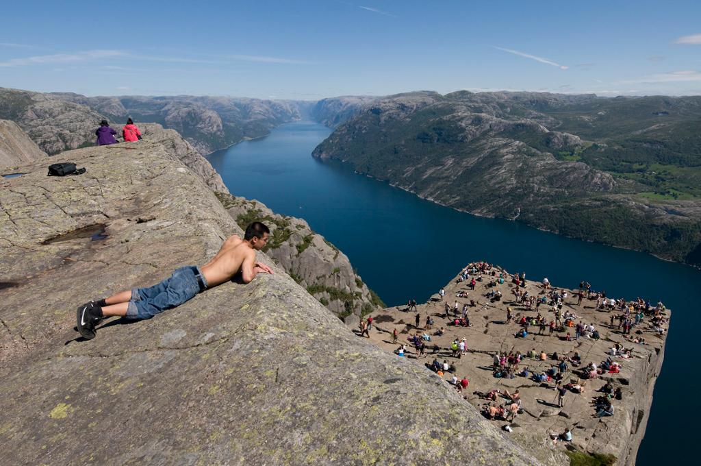 Ein gut ligg på eit berg og ser ut over fjorden. På ei berghylle lenger ned er det mange menneske. Foto.