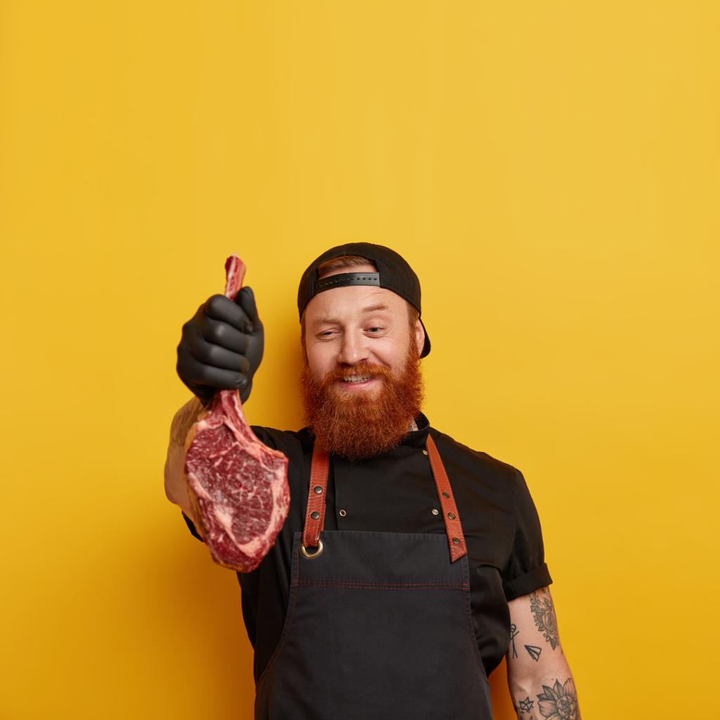 En kjøttskjærer viser fornøyd fram et rått kjøttstykke. Han har på seg svart arbeidstøy og svarte gummihansker. Foto.