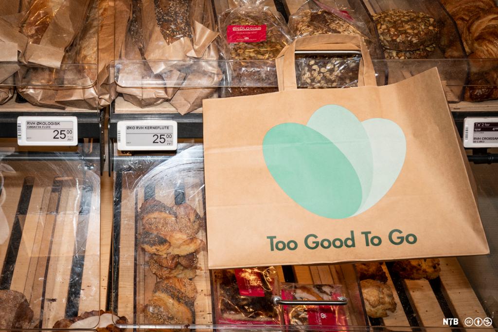 Flere typer brødvarer ligger i hyller med prislapp på hver enkelt vare. Noen av varene er innpakket i plast- eller papiremballasje. Over brødvarene ligger en bærepose i papir med logoen "Too Good To Go". Foto.