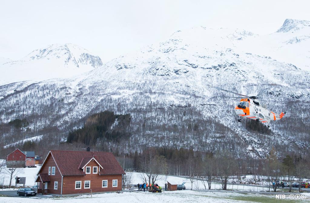 Eit helikopter i lufta og leitemannskapar på bakken ved eit hus nedanfor eit fjellparti. Foto.