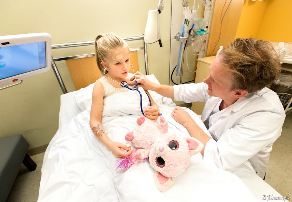 Ei jente med et plaster i panna sitter i ei seng på sykehuset. En stor, rosa bamse ligger på dyna foran henne. En lege sitter på huk ved senga og lytter på brystet hennes med stetoskop. Foto.
