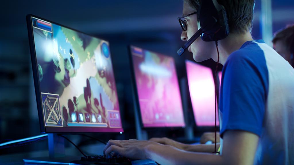 En gutt med headset sitter ved en PC og spiller et dataspill. Bak han er det flere skjermer og andre som spiller. Foto. 