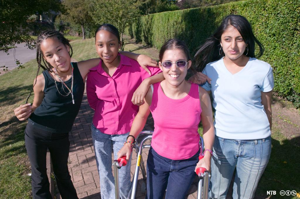 Jente med en funksjonsnedsettelse sammen med venninner. Jenta holder i en gåstol. Alle jentene holder rundt hverandre og smiler. Foto.