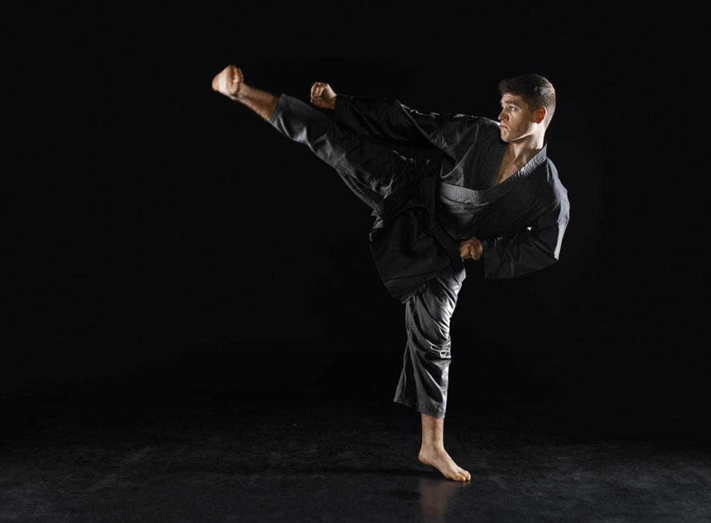 Mann med svart karatedrakt står på eitt bein og held det andre beinet svært høgt opp. Foto. 