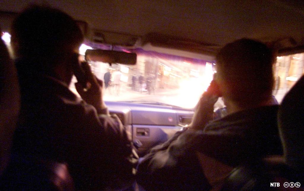 To personar, sett bakfrå, sit i ein bil mens dei kommuniserer og overvaker bylivet, som vi ser gjennom frontruta. Foto. 