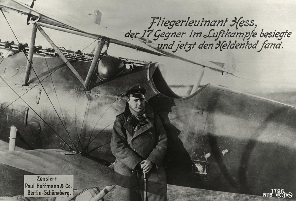 Mann med offiserslue og frakk foran en gammel flymodell. Bildet har teksten "flyløytnant Hess, som seiret 17 ganger i luftkamp, har nå lidd heltedøden". Foto.
