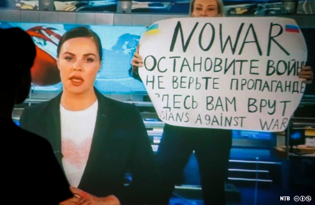 Eit kvinneleg nyheitsanker les nyheitsmeldingar på TV mens ei kvinne i bakgrunnen held opp ein plakat med bodskap på russisk og engelsk, kor det står "No war" og "Russians against war". Foto.