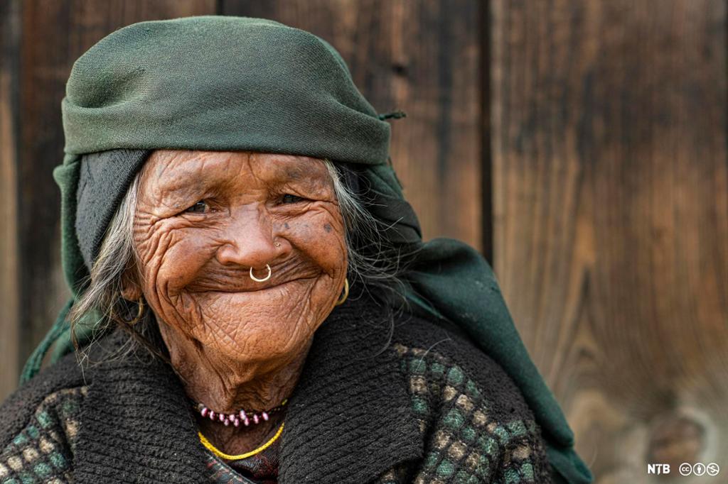Photo: We see an old woman. She has a lot of wrinkles. She is wearing necklaces and a nose ring. She is wearing a headscarf. She is wearing a wool jacket. She is smiling. From the photo description we know she is an old woman from the Himalayas.
