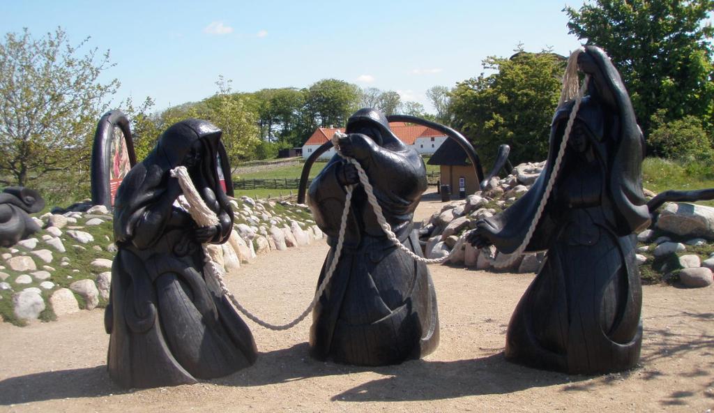 Statuer av tre svartkledde kvinner som held eit tau står på eit fast underlag. I bakgrunnen ser vi mark, tre og hus. Foto.