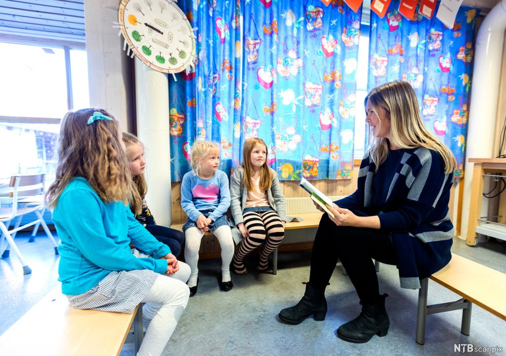 En voksen kvinne og fire jenter sitter sammen i et klasserom. Kvinnen leser fra ei bok. Elevene er i småskolealder. Foto.
