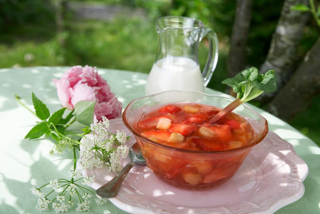 Jordbær- og rabarbrakompott i en glassbolle. Ved siden av står en mugge med fløte. Desserten står på et bord utendørs, og bordet er også pyntet med blomster. Foto.