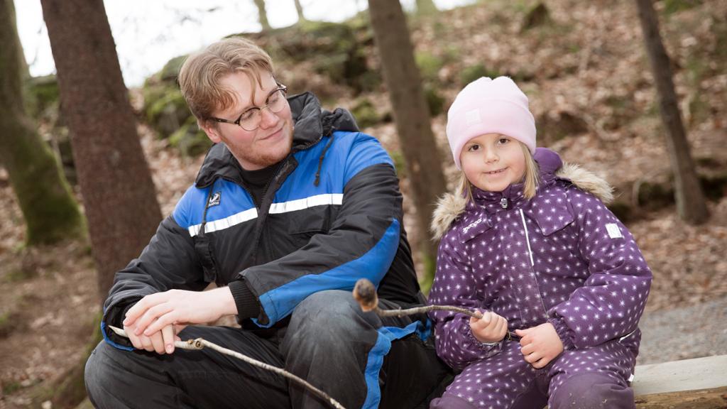 Jente i lilla utedress sitter med et svidd pinnebrød sammen med en voksen på en benk. Foto.