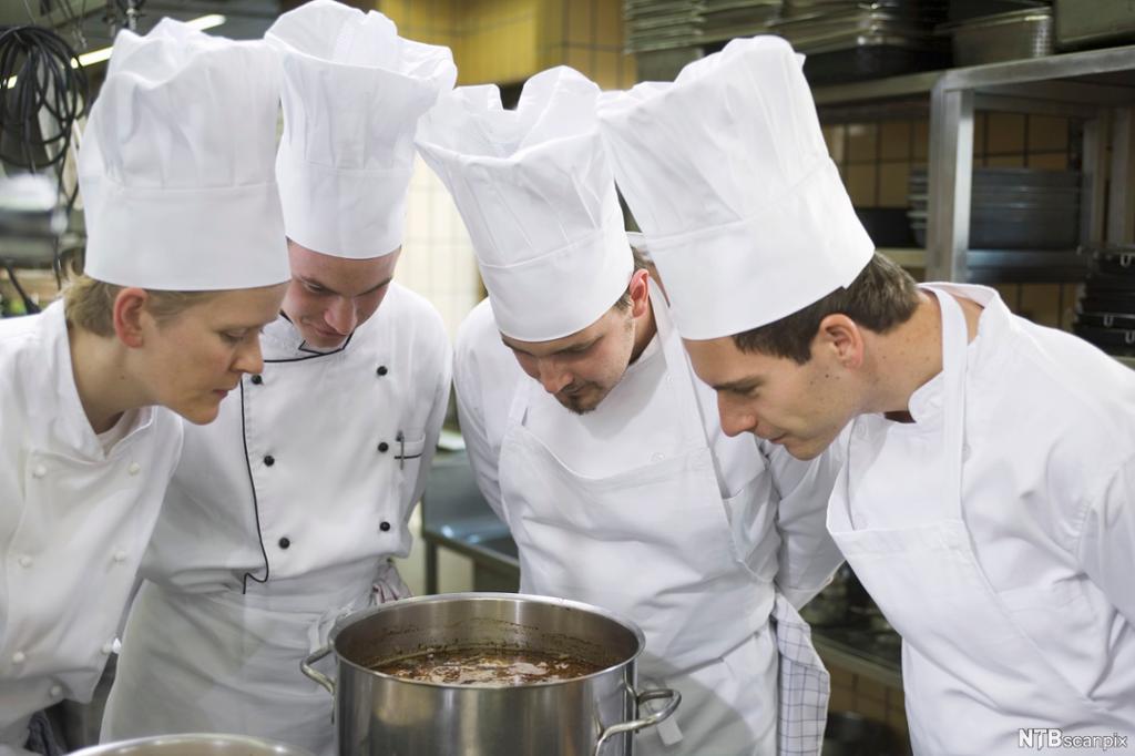 Fire mannlege kokkar i kokkeuniform står med hendene på ryggen og ser ned i ein stor kjele. Foto.