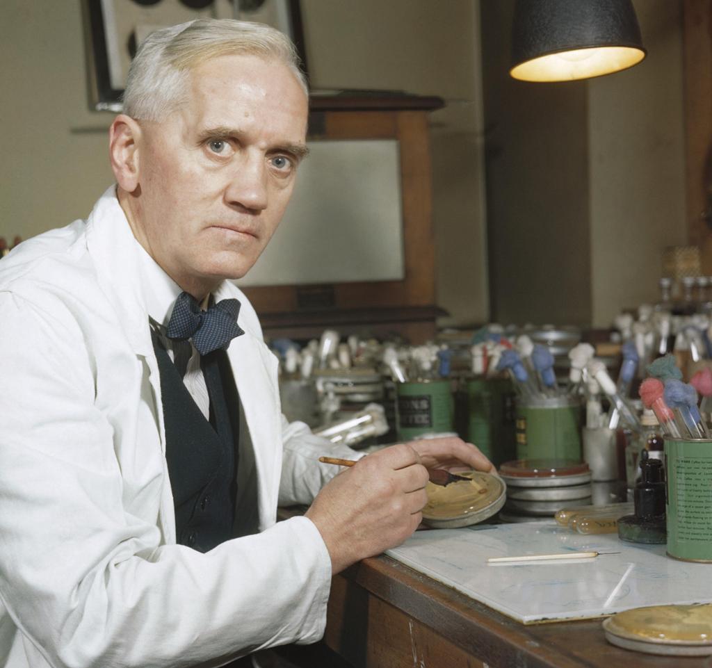 En mann sitter i hvit frakk på et laboratorium og undersøker ei agarskål med penicillin. Foto. 