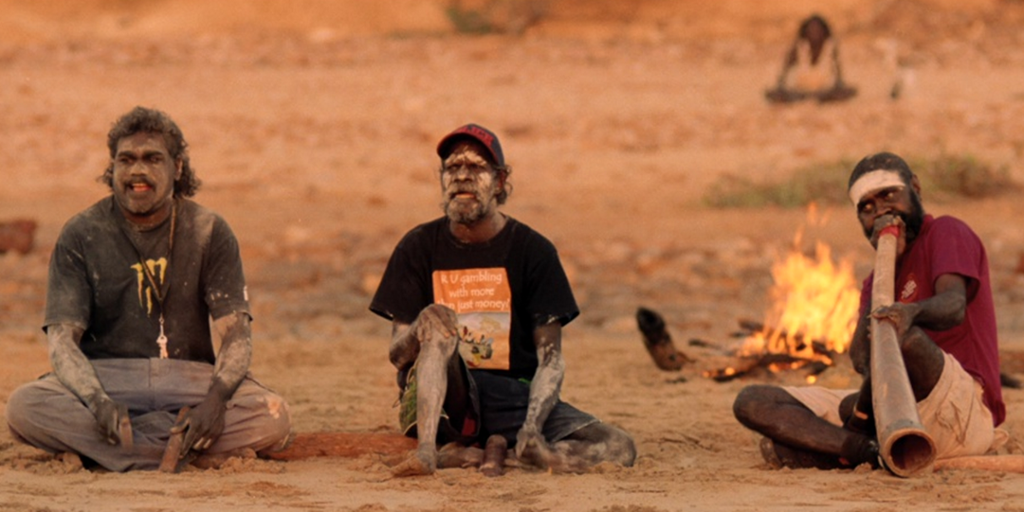 Tre aboriginske menn sitter og spiller tradisjonelle didgeridoo- og slaginstrumenter foran et bål i den australske ørkenen. Foto.