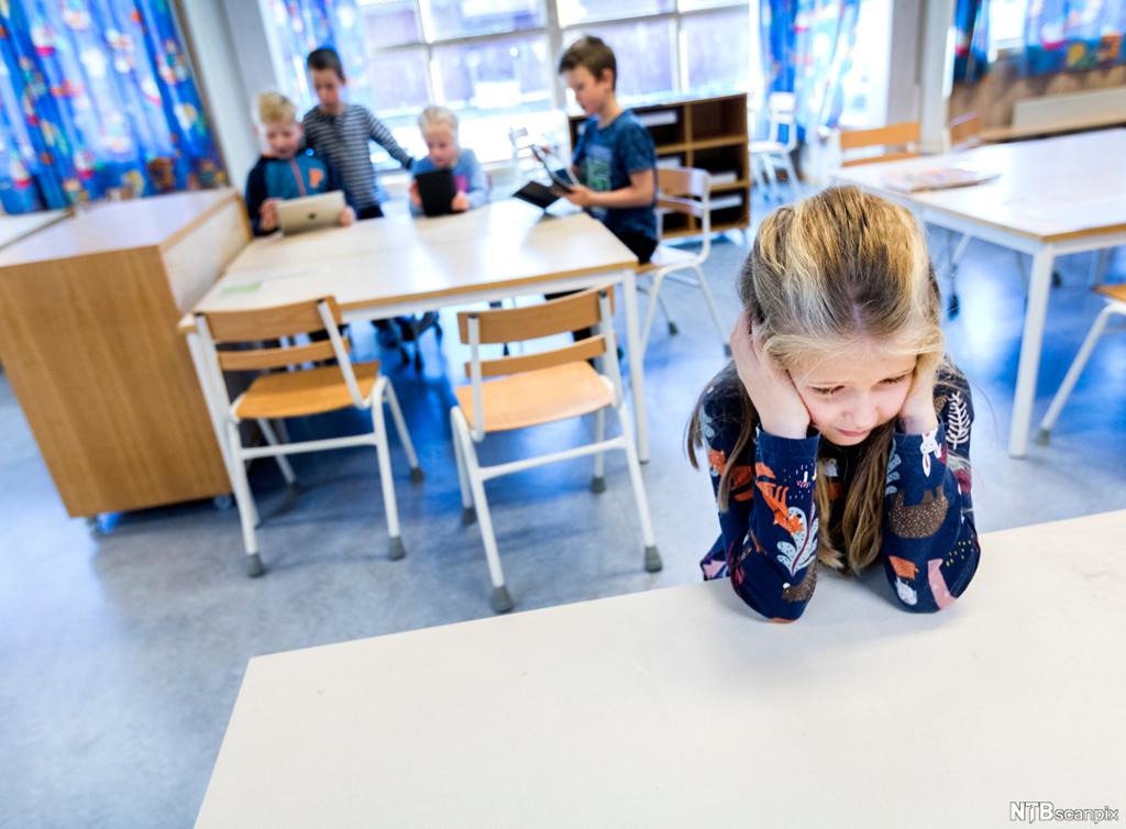 Ei jente sitter alene ved en pult i et klasserom. Hun støtter hodet i hendene og ser trist ut. I bakgrunnen sitter fire andre barn ved et bord. Foto.
