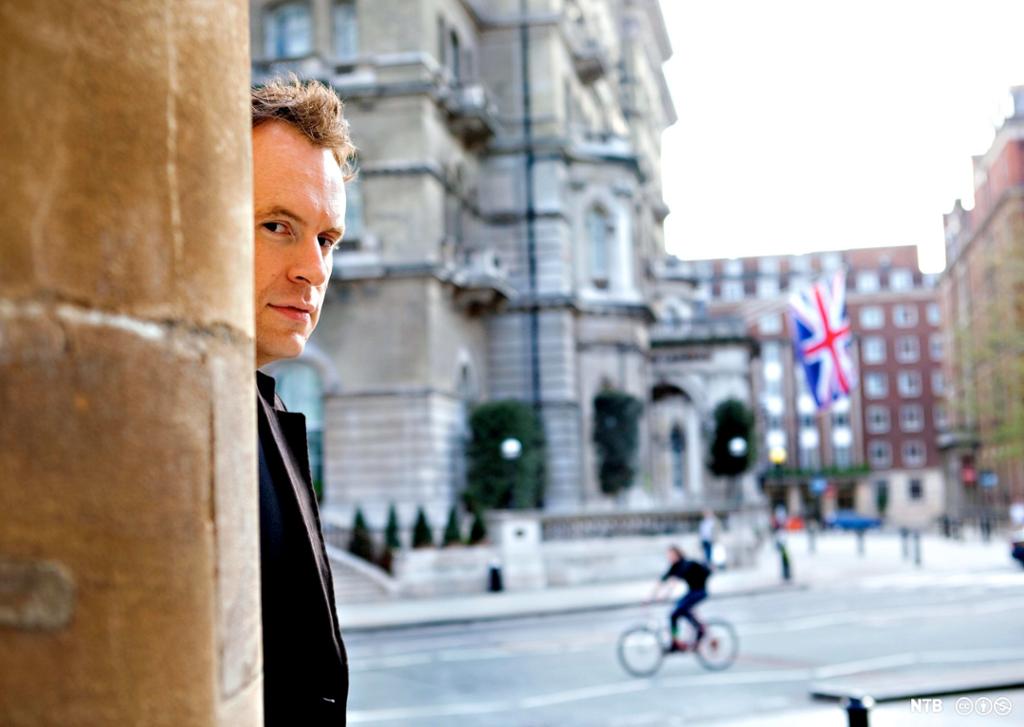 Matt Haig looking from behind a wall, a street and a Union Jack in the background. 