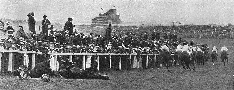 An old black and white photo from the Epsom Derby. Suffragette Emily Davison has just flung herself in front of a horse, and the horse, the jockey, and Ms Davison are all down. There are many people watching the race and we see the back of the remaining horses as they continue the race.